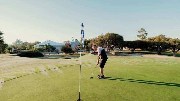 Un terrain de golf avec un mât de drapeau, un golfeur portant un t-shirt blanc et un short noir, et un bâtiment en arrière-plan.
