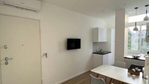 Une cuisine moderne avec des meubles blancs, un téléviseur noir fixé au mur et un coin repas comprenant une table blanche et deux chaises, une jaune et une grise.