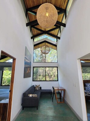 A spacious room with a high ceiling, a woven light fixture, a large window with a view of trees, a dark-colored sofa, and a small wooden table.