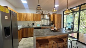 A modern kitchen with wooden cabinets, a central island with stools, pendant lighting, and a large window with a view of greenery. - Casa Vail, surrounded by jungle in Nosara area (Nosara)