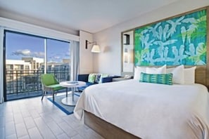 Une chambre d’hôtel avec un grand lit, un balcon donnant sur la ville, une petite table avec une chaise et un tableau au mur.