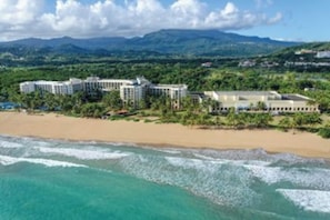 Un hôtel situé en bord de mer, composé de plusieurs bâtiments, avec une plage de sable et une vue sur les montagnes lointaines.