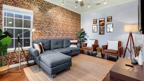 A living room with a brick wall, a sectional sofa, a brown leather chair, a wooden coffee table, and a floor lamp. - Spacious Retreat & Game Room Sleeps 12 (New Braunfels)