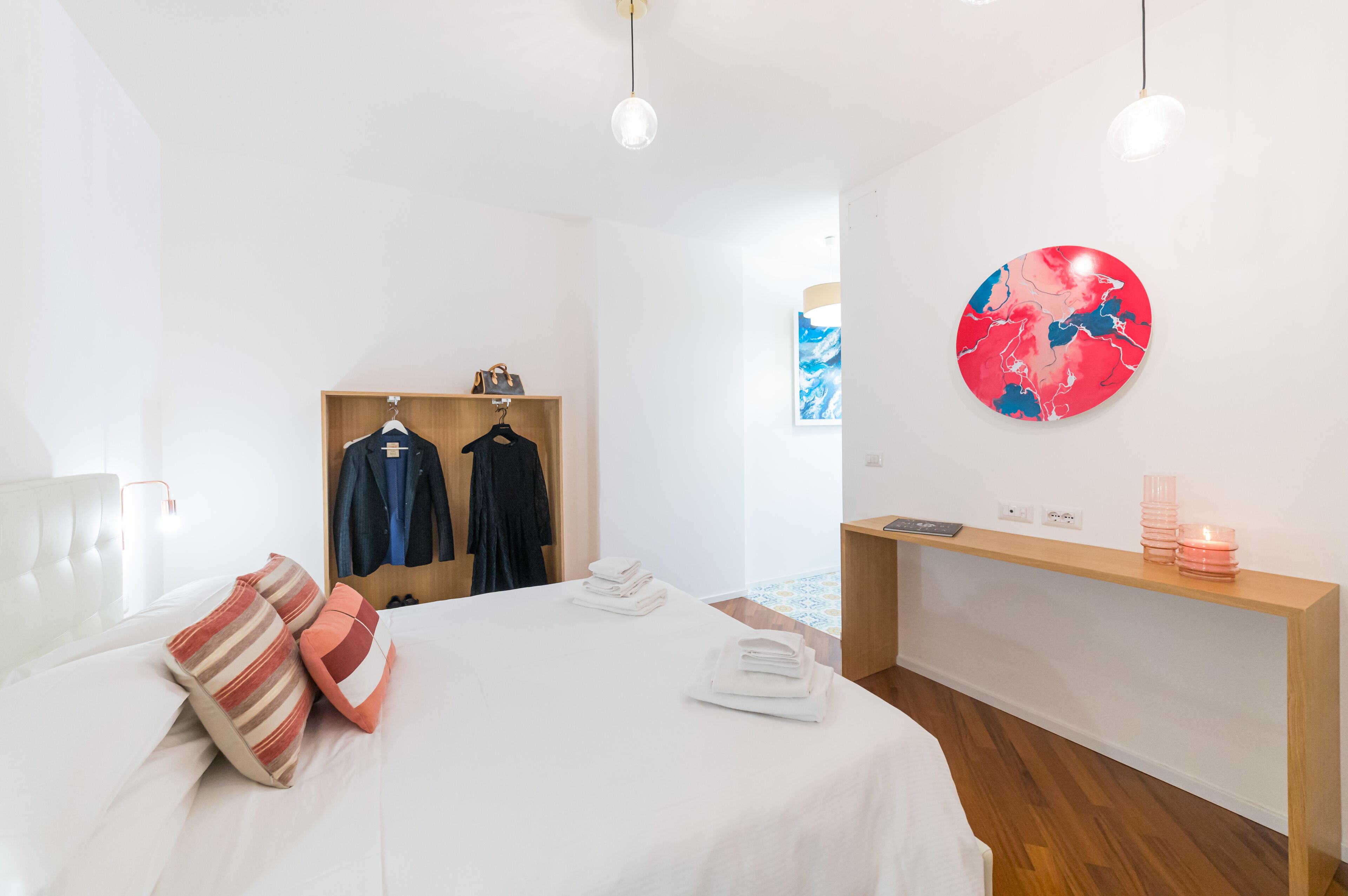 A modern hotel room with a neatly made bed, a wooden wardrobe, a round artwork on the wall, and a small wooden side table with a lamp.