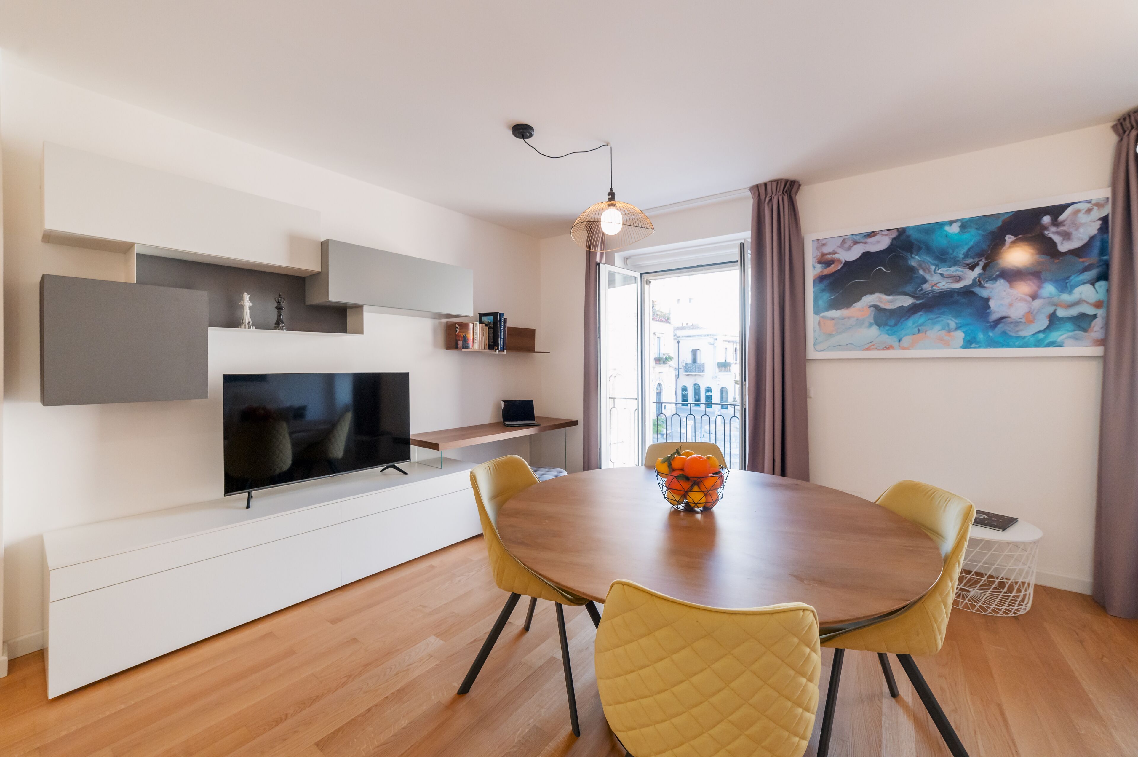 Una sala de estar moderna con un televisor de pantalla plana, una mesa de comedor con sillas, un frutero y un cuadro abstracto grande.