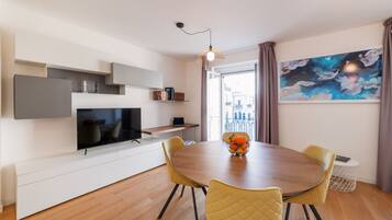 Una sala de estar moderna con un televisor de pantalla plana, una mesa de comedor con sillas, un frutero y un cuadro abstracto grande.