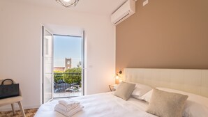 A hotel room with a large bed, a nightstand with a lamp, a chair, and a view of a building with a clock tower through a glass door. - Castalia Apartments & Rooms (Taormina)