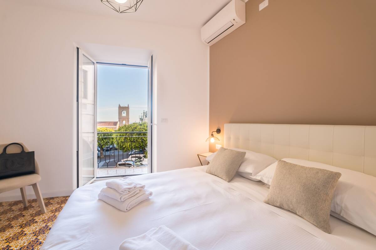 A hotel room with a large bed, a nightstand with a lamp, a chair, and a view of a building with a clock tower through a glass door.