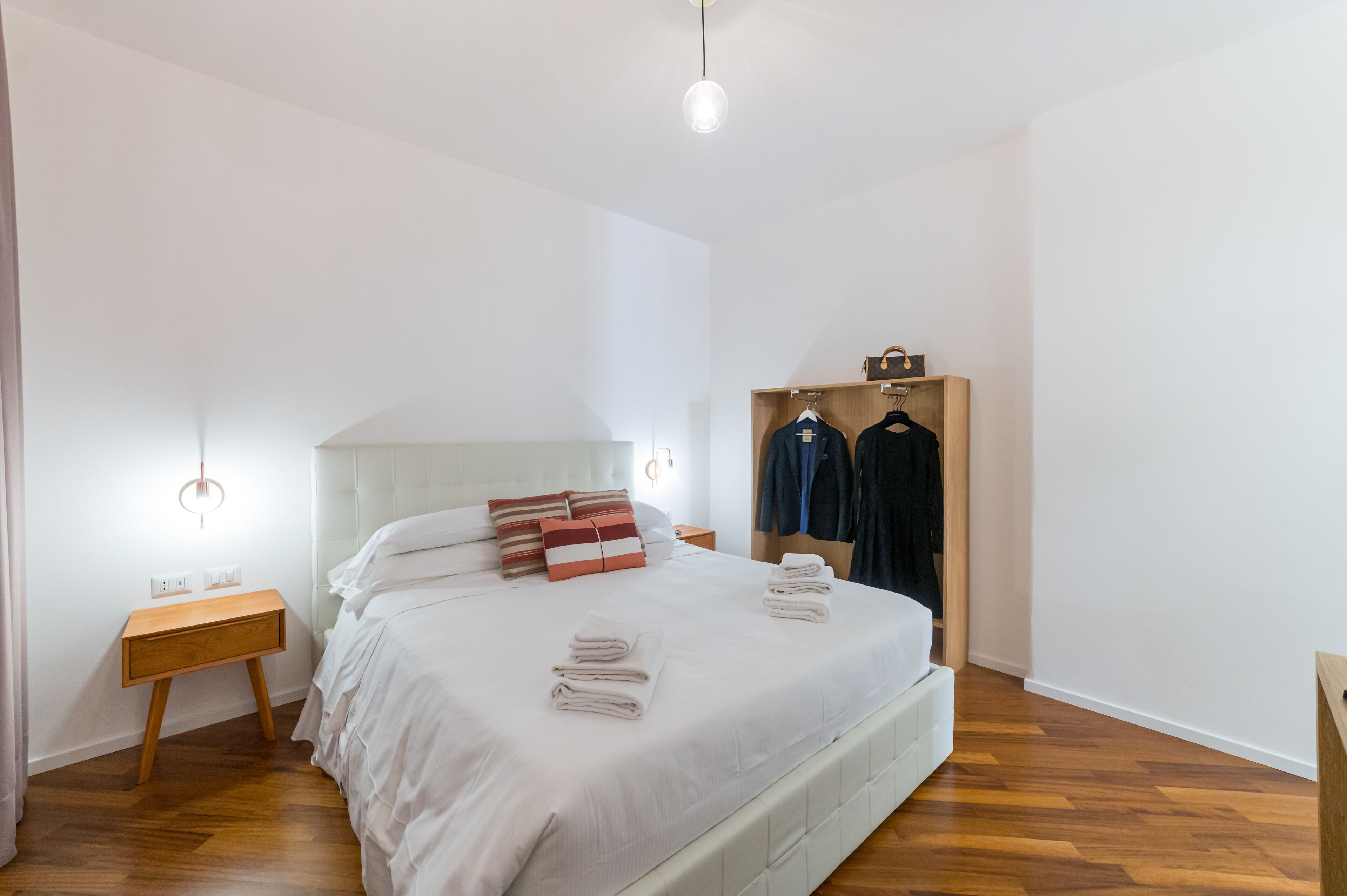 A neatly made bed with white linens, a wooden nightstand, a wooden wardrobe, and a hanging light fixture.