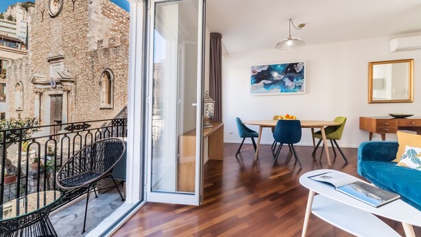 A modern living room with a balcony, wooden flooring, and a dining area with chairs and a table. - Castalia Apartments & Rooms (Taormina)