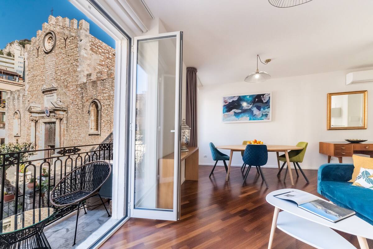A modern living room with a balcony, wooden flooring, and a dining area with chairs and a table.