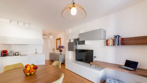 A modern kitchen with white cabinets, a dining table with a fruit bowl, a TV, and a laptop on a wooden shelf. - Castalia Apartments & Rooms (Taormina)