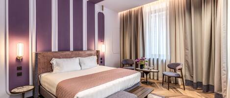 A modern hotel room with a large bed, a small table, two chairs, and a large window with curtains.