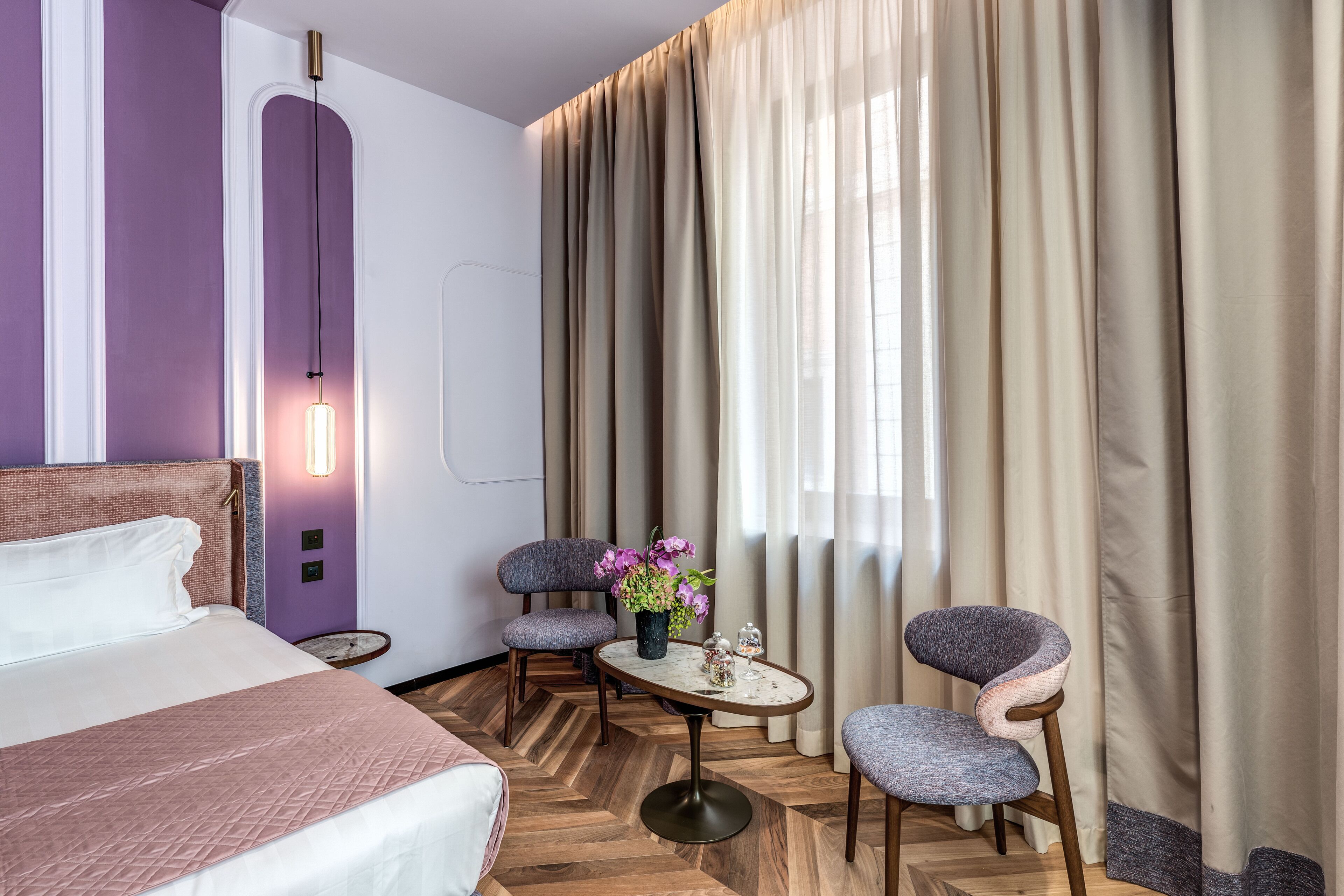 A hotel room with a bed, two chairs, a small table with flowers, and a large window with curtains.