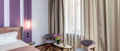 A hotel room with a bed, two chairs, a small table with flowers, and a large window with curtains.