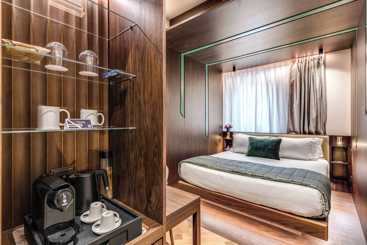 Habitación de hotel moderna con pared de paneles de madera, cama con un plaid verde, estación de café y ventana con cortinas.