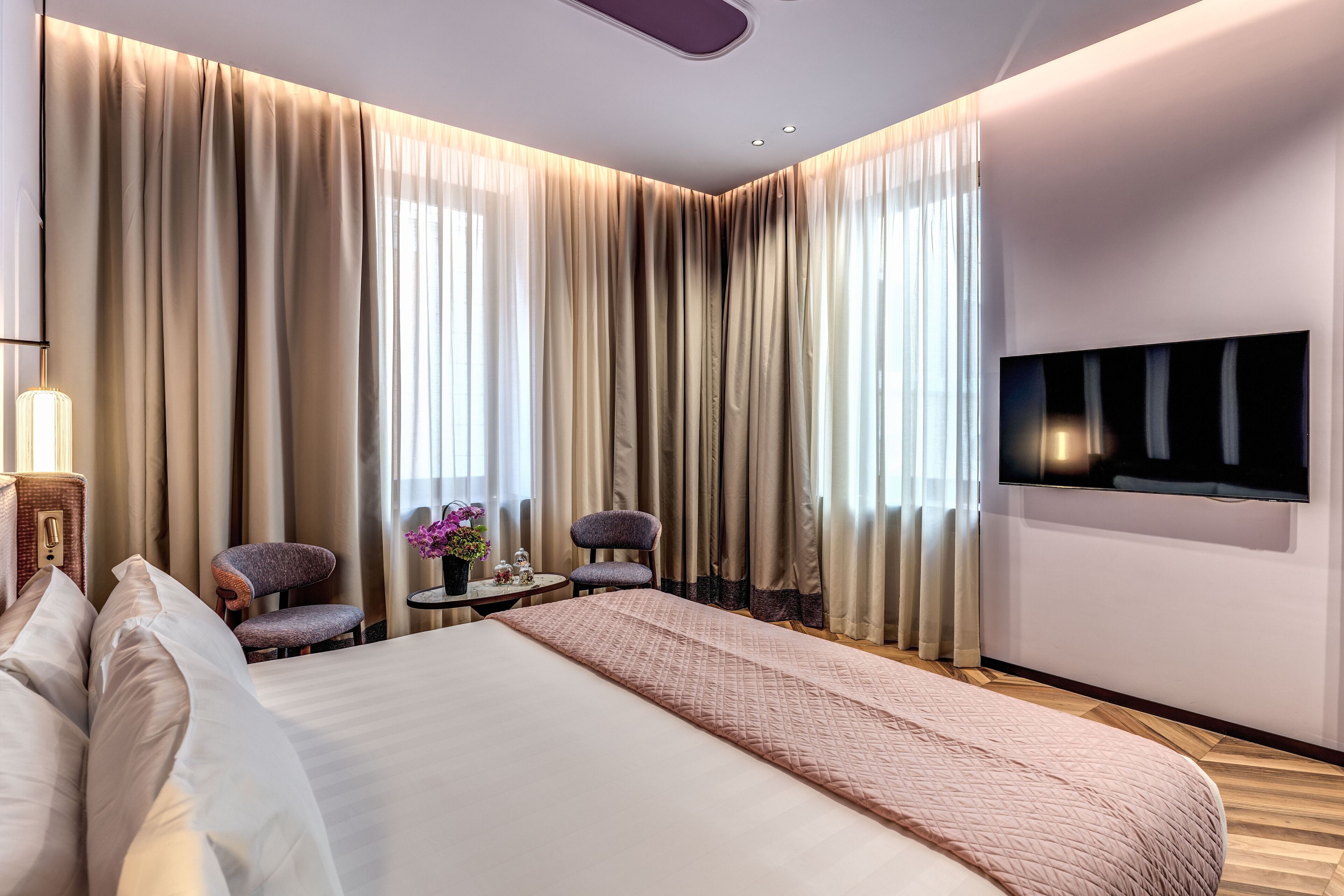 A modern hotel room with a large bed, a flat-screen TV mounted on the wall, a small table with a vase of flowers, and two chairs.
