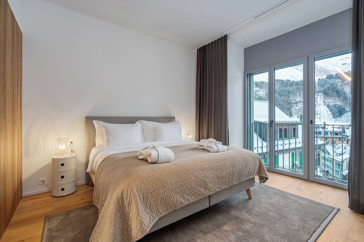 A modern bedroom with a large bed, a bedside table with a lamp, and a view of a snowy landscape through large windows.
