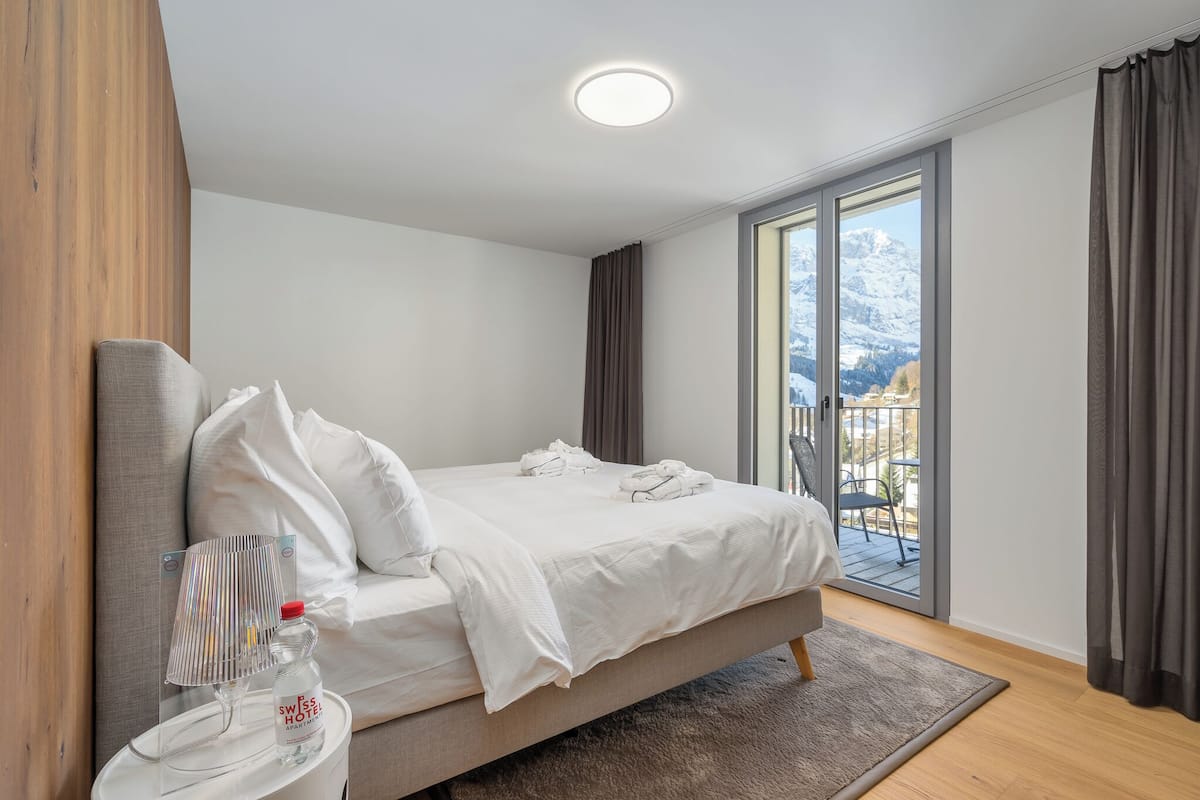A modern hotel room with a large bed, a small side table, a bottle of water, and a view of a snowy mountain through a sliding glass door.
