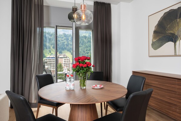 Un espace repas moderne avec une table ronde en bois, des chaises noires, un vase rempli de fleurs rouges et une vue sur un paysage montagneux grâce à de grandes fenêtres.