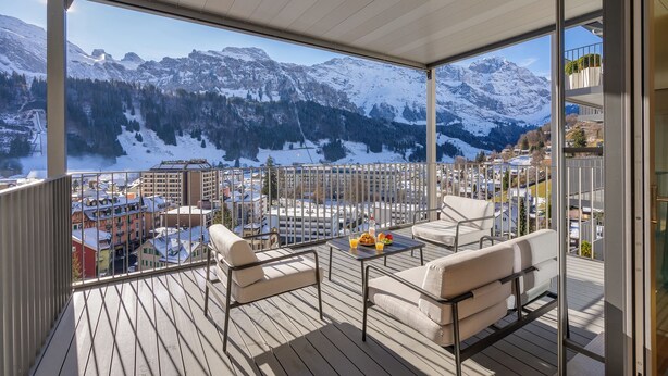 A balcony with a view of snow-covered mountains, a table with a fruit bowl, and two sofas.