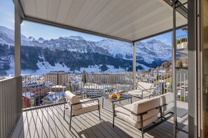 Un balcon avec vue sur des montagnes enneigées, une table avec un panier de fruits et deux canapés.