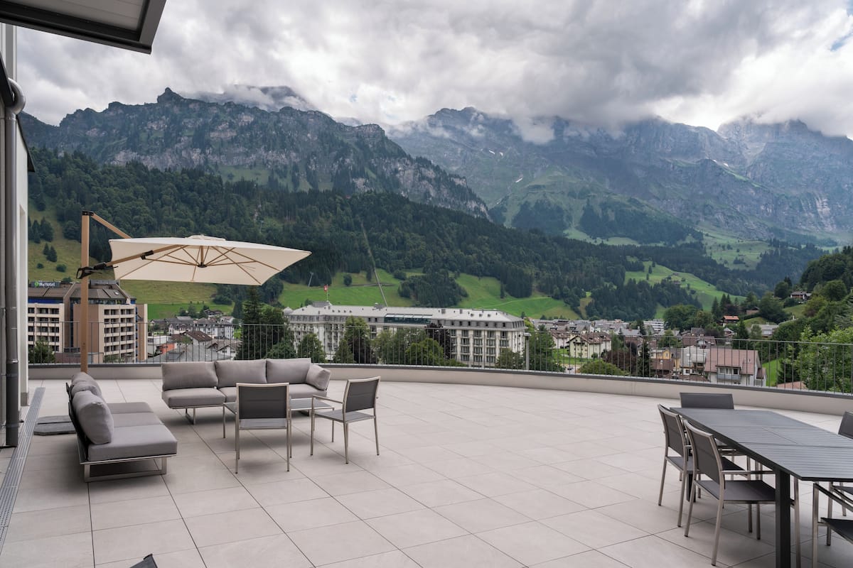 A terrace with outdoor furniture, a table, and an umbrella, overlooking a mountainous landscape.