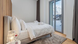 A modern bedroom with a large bed, a nightstand, a lamp, and a view of a snowy mountain through a sliding glass door.