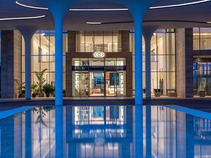 Une entrée d’hôtel moderne avec de grandes portes vitrées, une piscine au premier plan et le reflet du bâtiment.