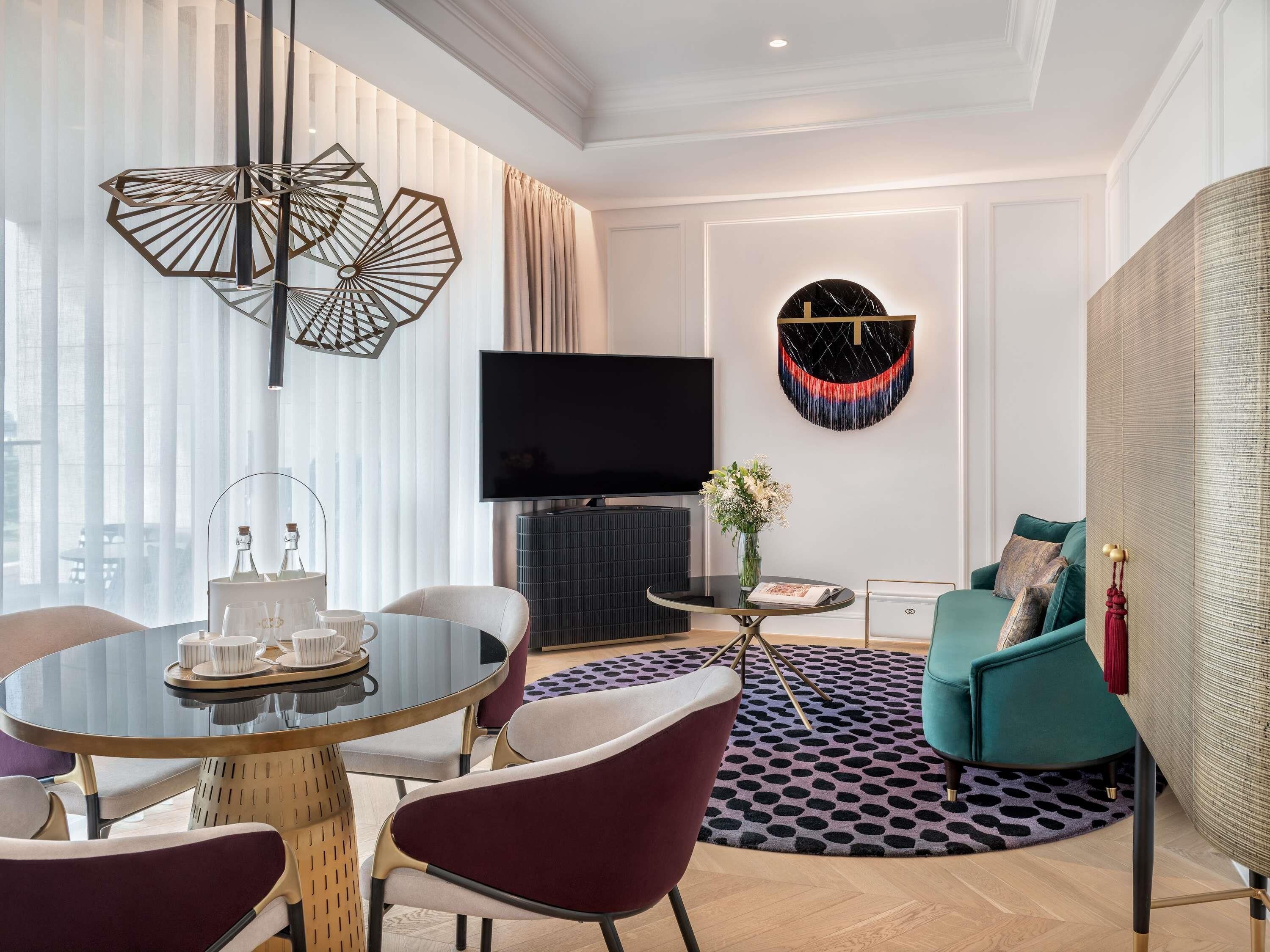 A modern living room with a round glass table, red chairs, a flat-screen TV, a decorative wall piece, and a patterned rug.