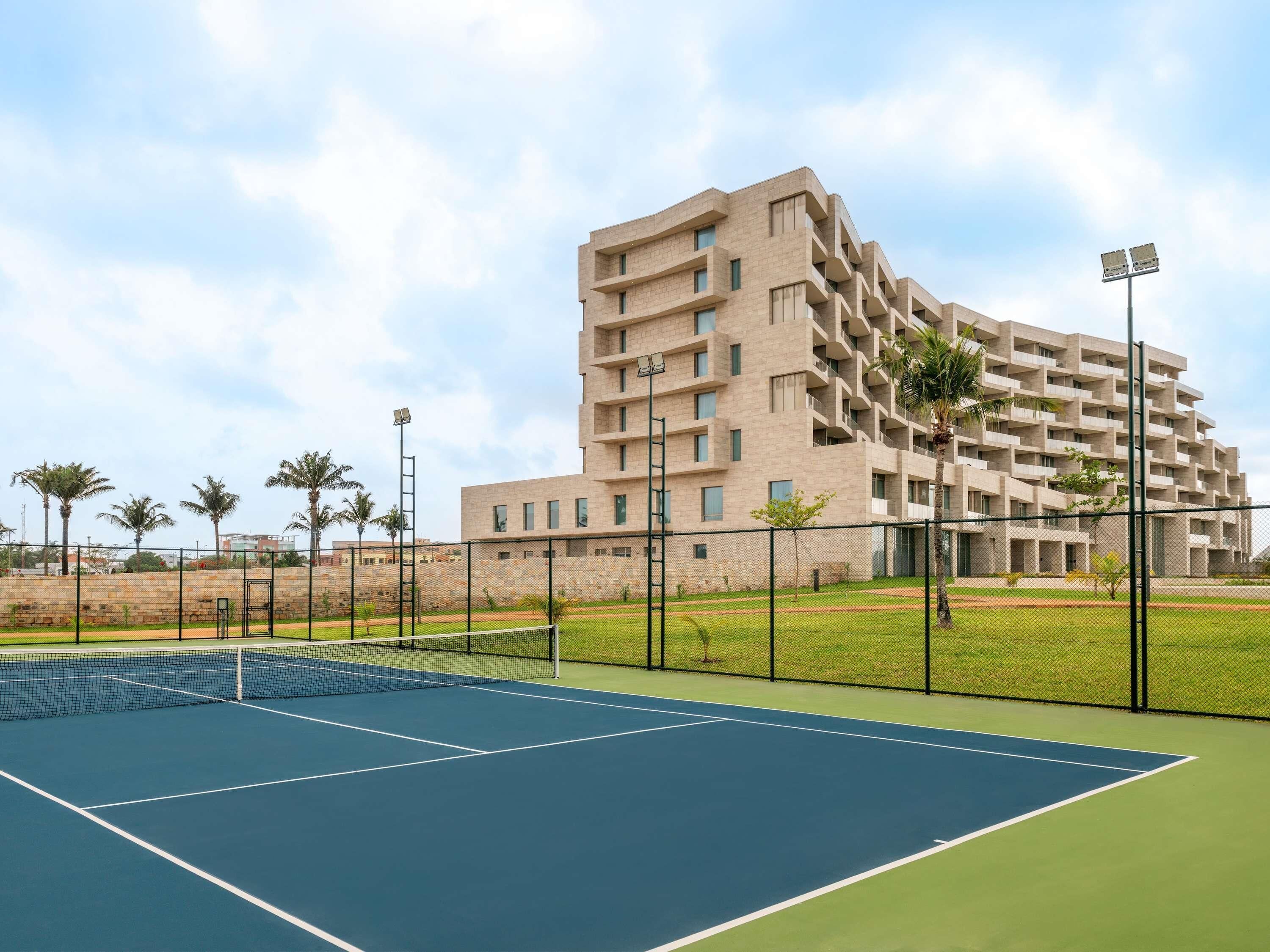 A tennis court with a net, surrounded by a fence, and a modern building with multiple balconies in the background.