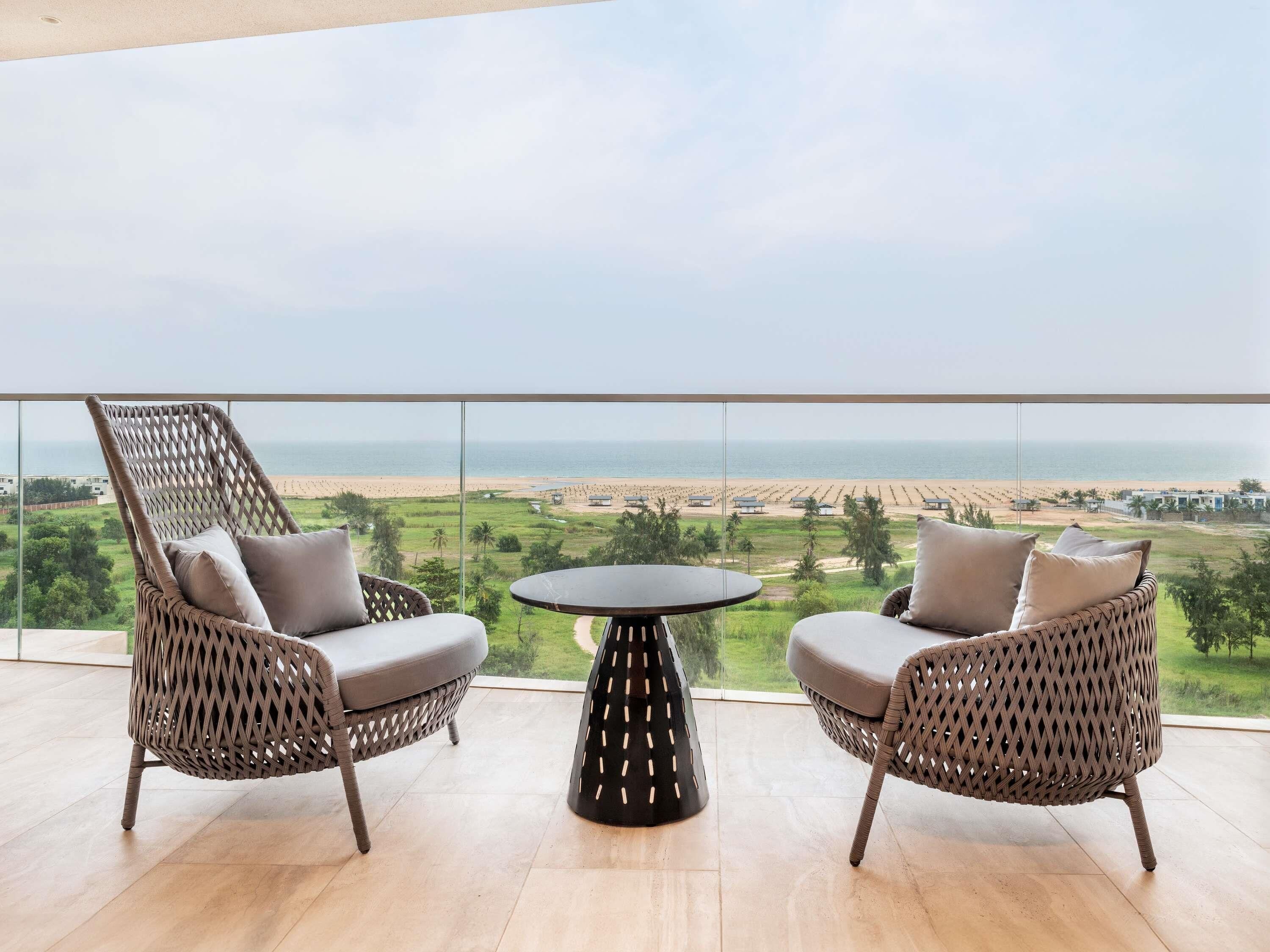 A balcony with wicker chairs and a table, overlooking a beach and greenery.