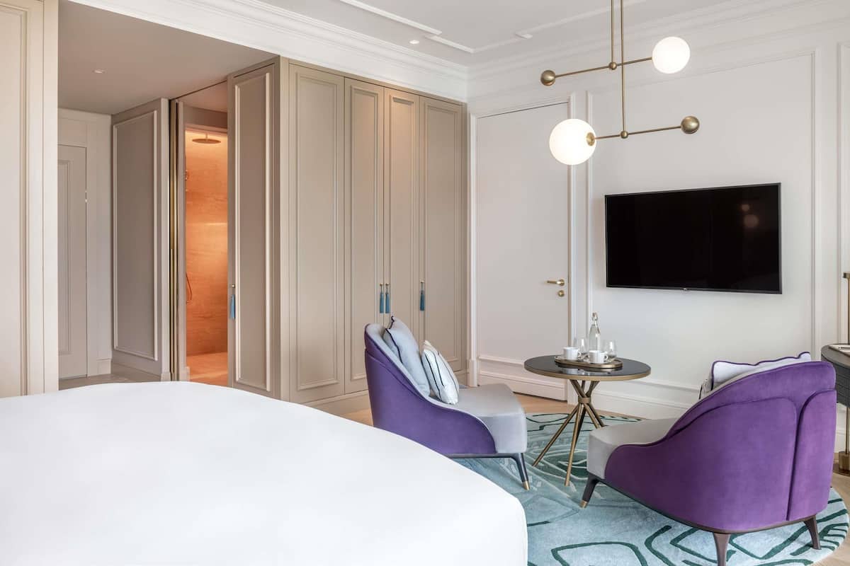Een hotelkamer met een groot bed, twee paarse fauteuils, een ronde tafel, een aan de muur bevestigde televisie en een garderobe met spiegel.