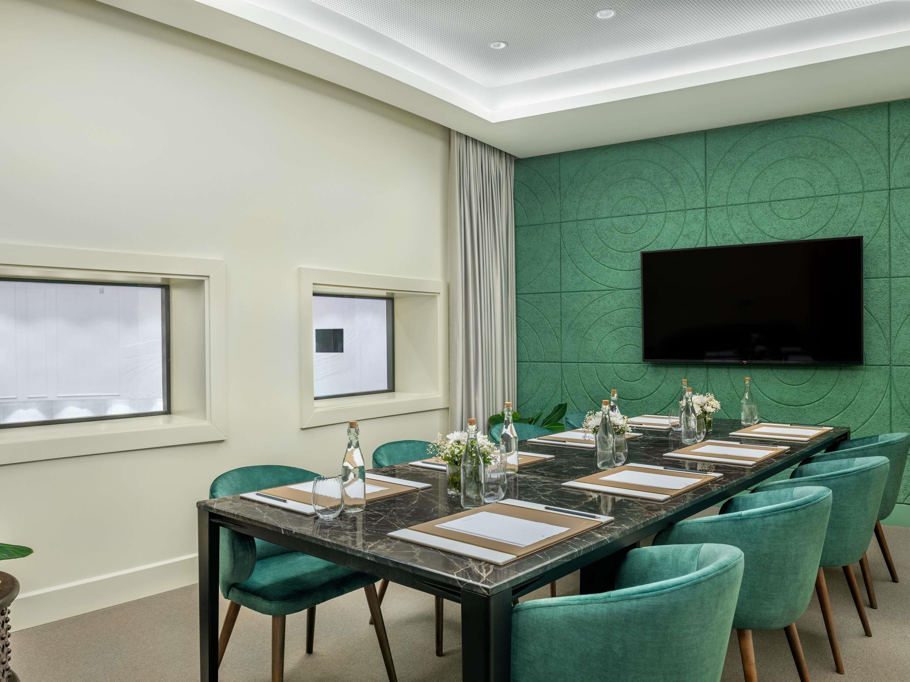 A conference room with a long table, teal chairs, a flat-screen TV, and a green textured wall.