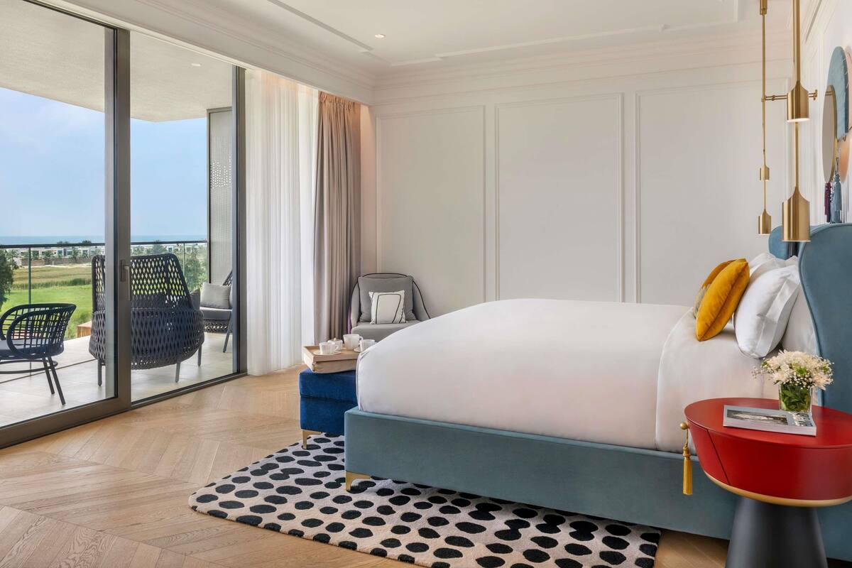 Een slaapkamer met een groot bed, een rode bijzettafel, een stoel, een tafeltje met kopjes, en uitzicht door een glazen deur.