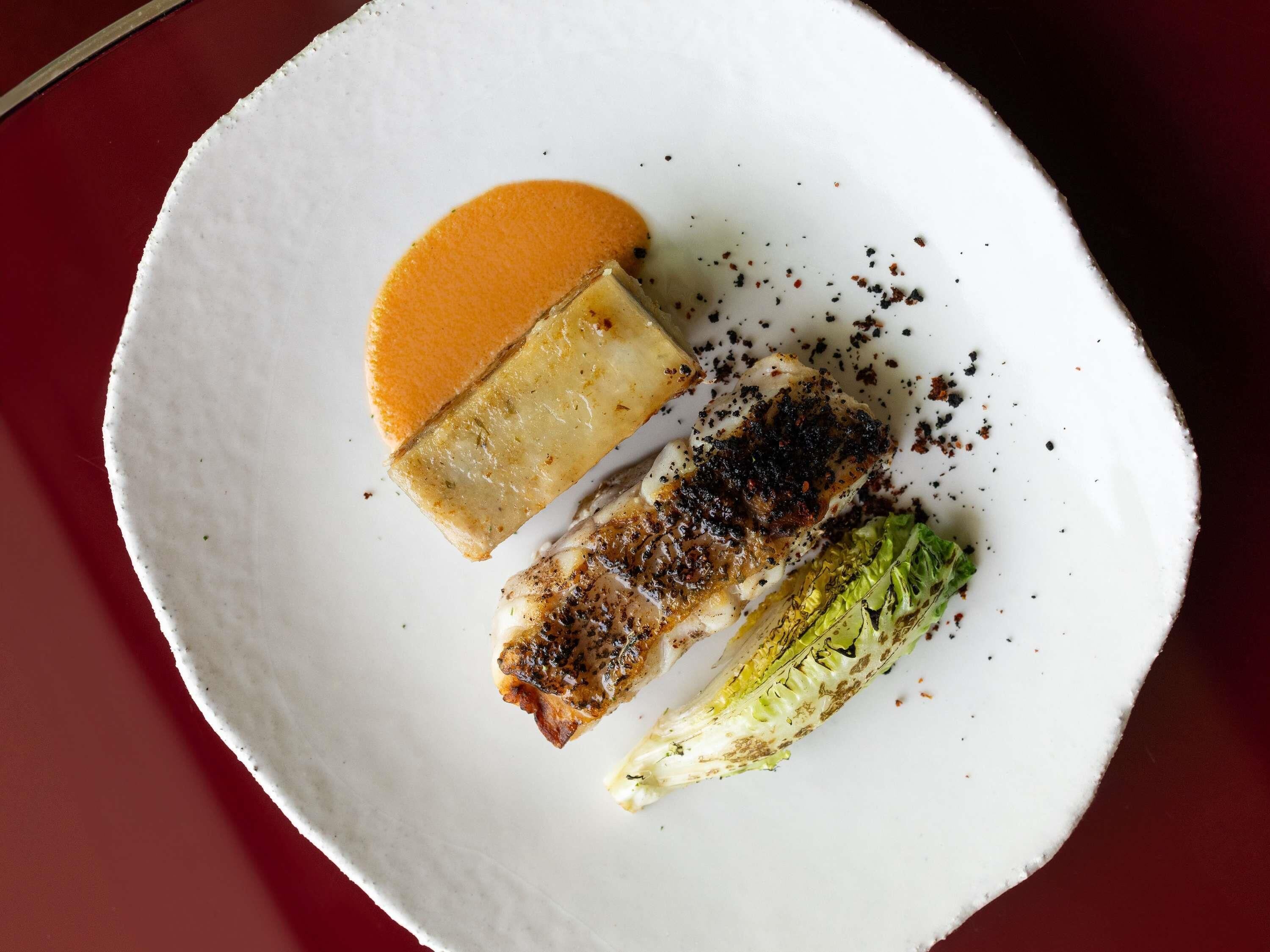 Un plato blanco con un trozo de pescado a la parrilla, una porción de queso y una pequeña cantidad de lechuga.