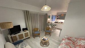 Una sala de estar moderna con un sofá, dos sillones, una mesa de centro, una televisión y una cocina al fondo.