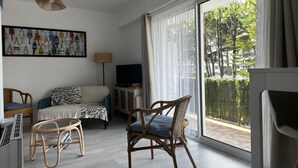 Una sala de estar moderna con un sofá, dos sillones de mimbre, un televisor y un ventanal con vistas a árboles.