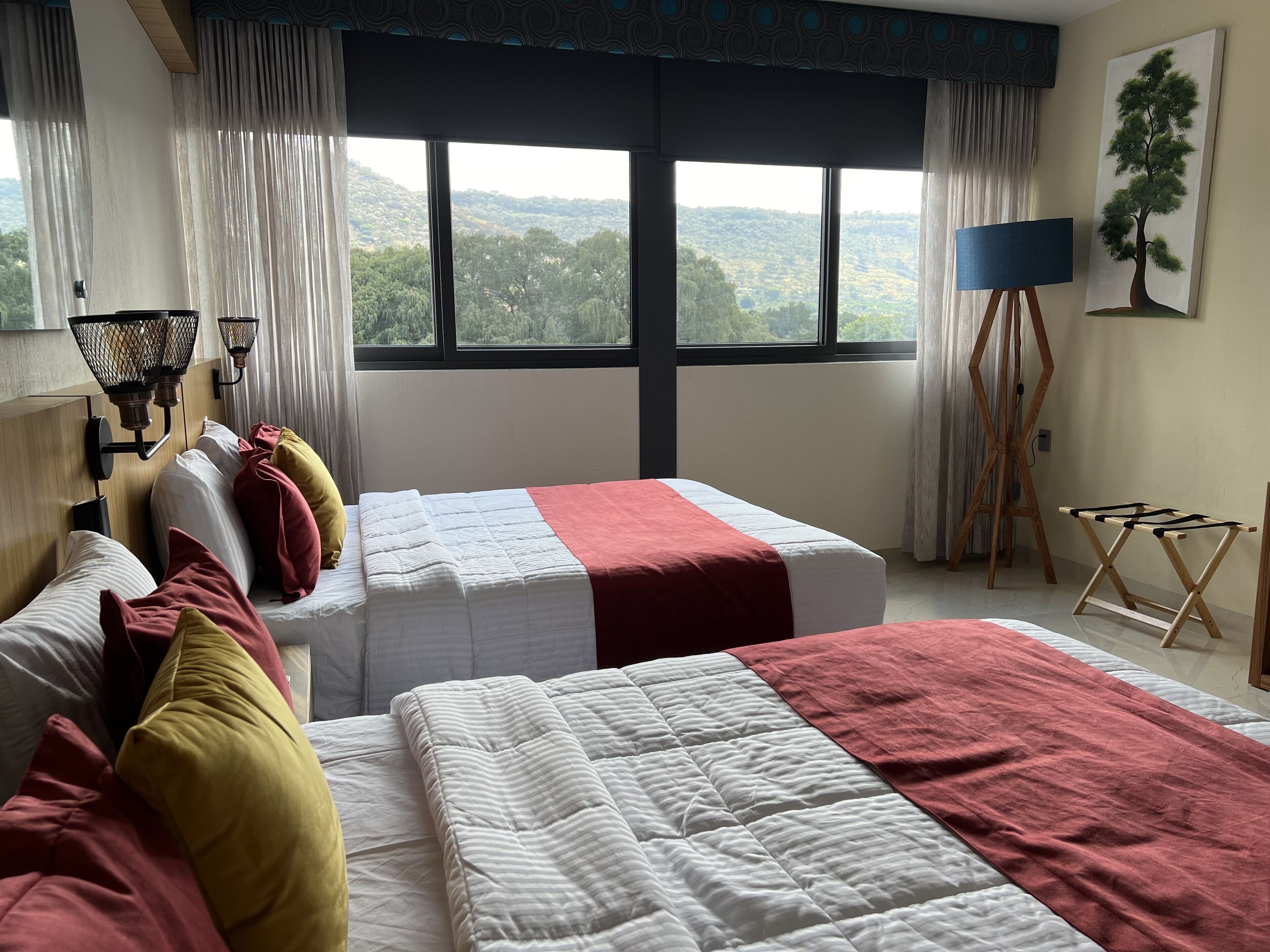 A hotel room with two beds, a large window with a view of the countryside, a standing lamp, and a painting on the wall.