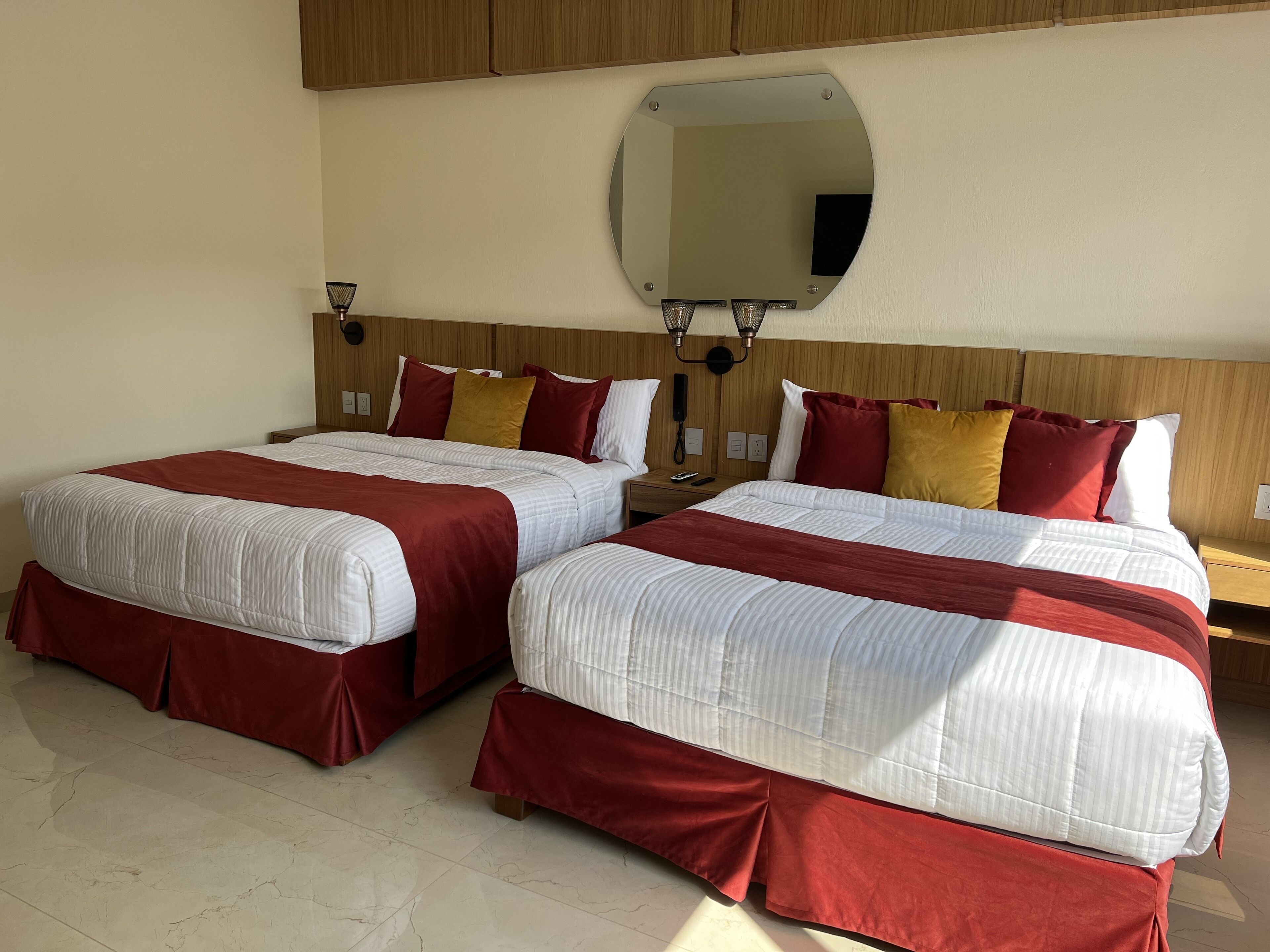 A hotel room with two beds, each with red and white bedding, a wooden headboard, a round mirror, and a bedside lamp.