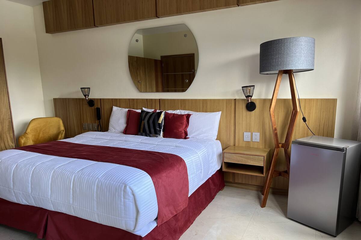 Habitación de hotel con una cama grande, cabecera de madera, lámparas de noche, una mesita pequeña y un refrigerador compacto.