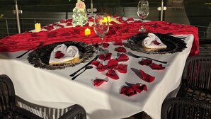 Mesa de comedor al aire libre decorada con pétalos de rosa roja, velas y un jarrón de rosas.