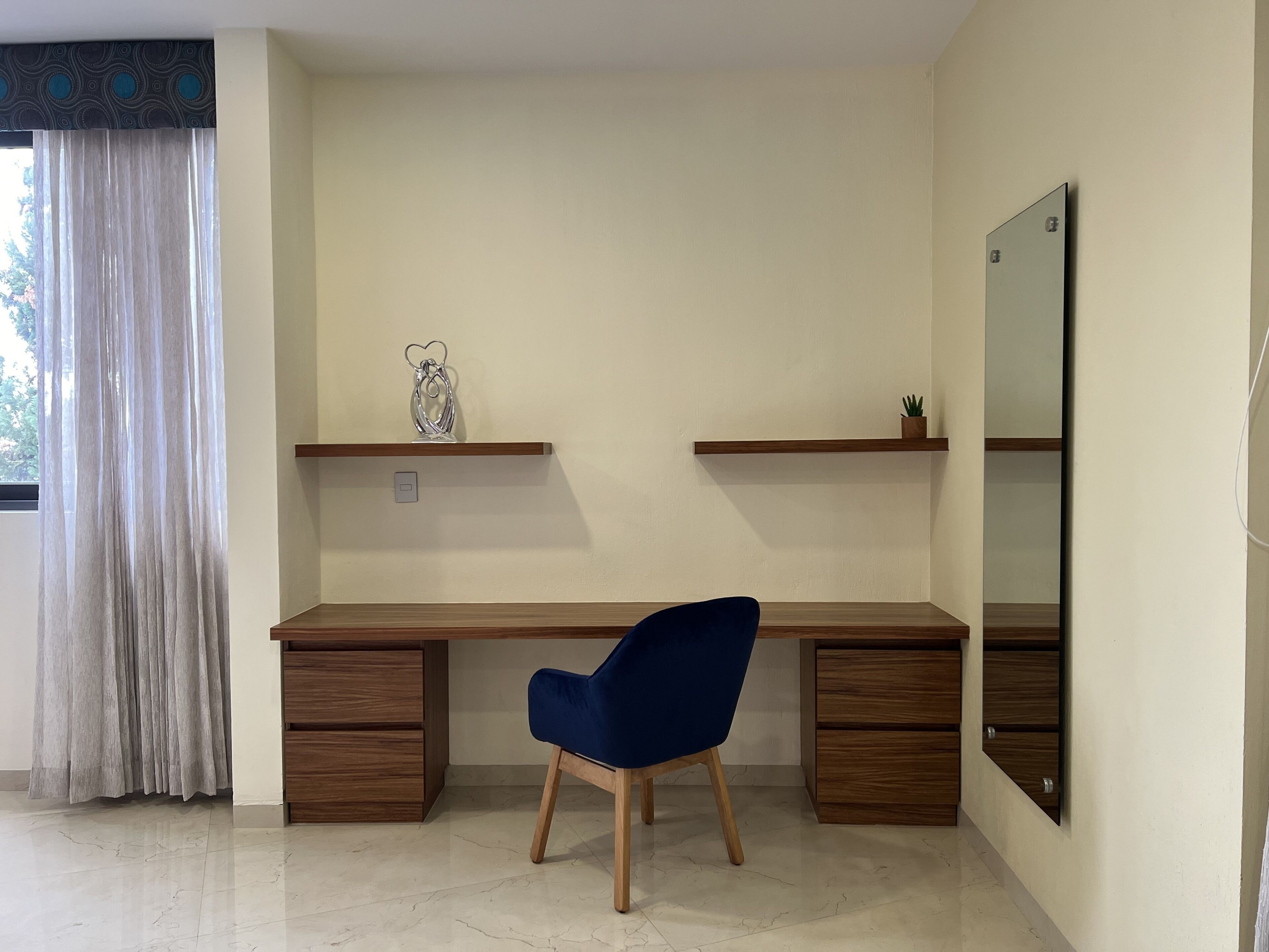 A modern study room with a wooden desk, two drawers, a blue chair, a large mirror, and a window with curtains.