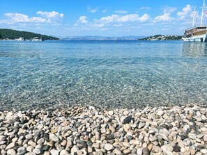 A pebbly beach with clear water, boats, and a sailboat in the distance.