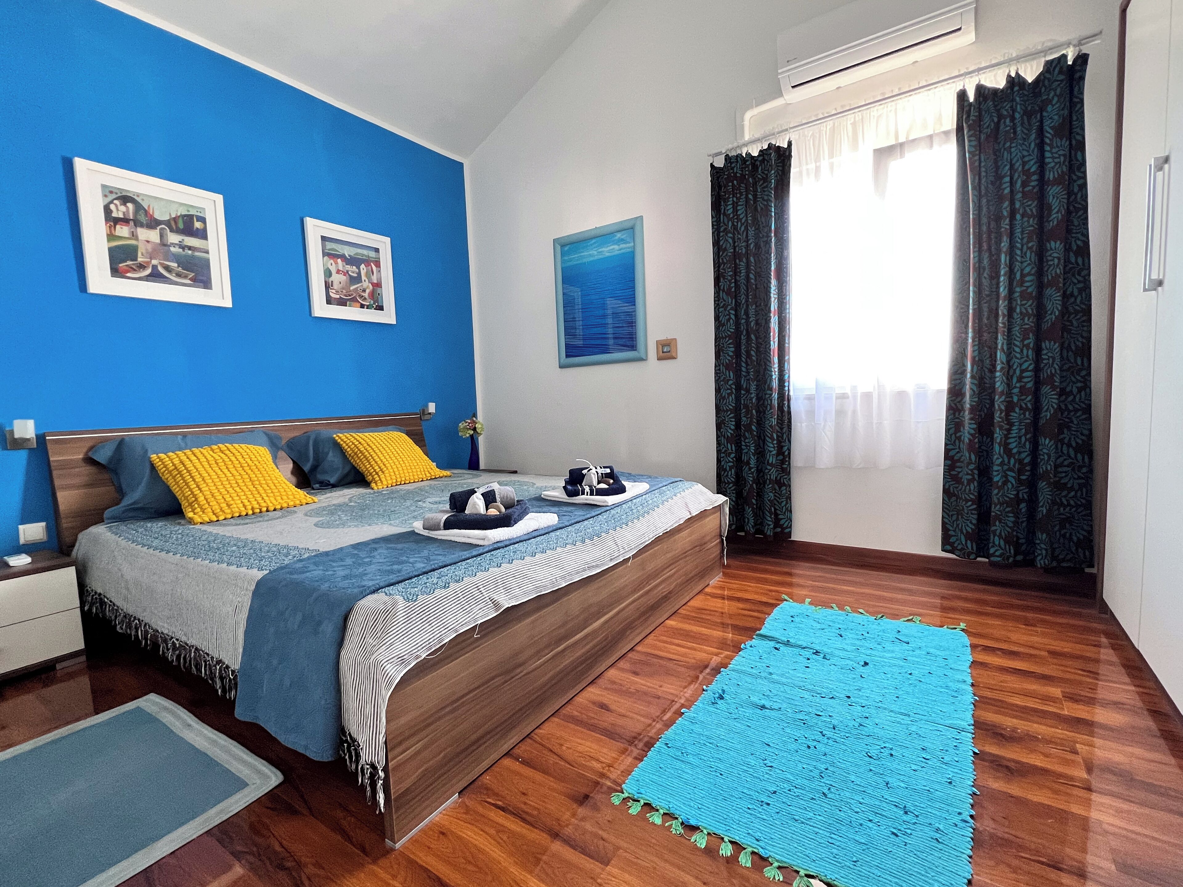 Quarto com cama de madeira, paredes azuis e uma janela grande com cortinas.