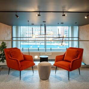 A modern lobby with two orange chairs, a grey ottoman, and a large window with a view of buildings. - Aparthotel Birmingham (BIRMINGHAM)
