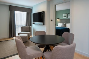 A hotel room with a round table, four chairs, a TV, a bed, and a view of the outside.