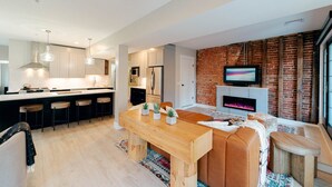 A modern kitchen with a central island, bar stools, and a dining area with a wooden table and seating. A fireplace and TV are visible in the living area.