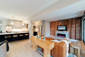 Een moderne keuken met een centraal eiland, barkrukken en een eethoek met een houten tafel en zitplaatsen. In de woonkamer is een open haard en een televisie te zien.