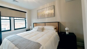 A bedroom with a large bed, a window with blinds, a wall-mounted artwork, and a nightstand with a lamp.
